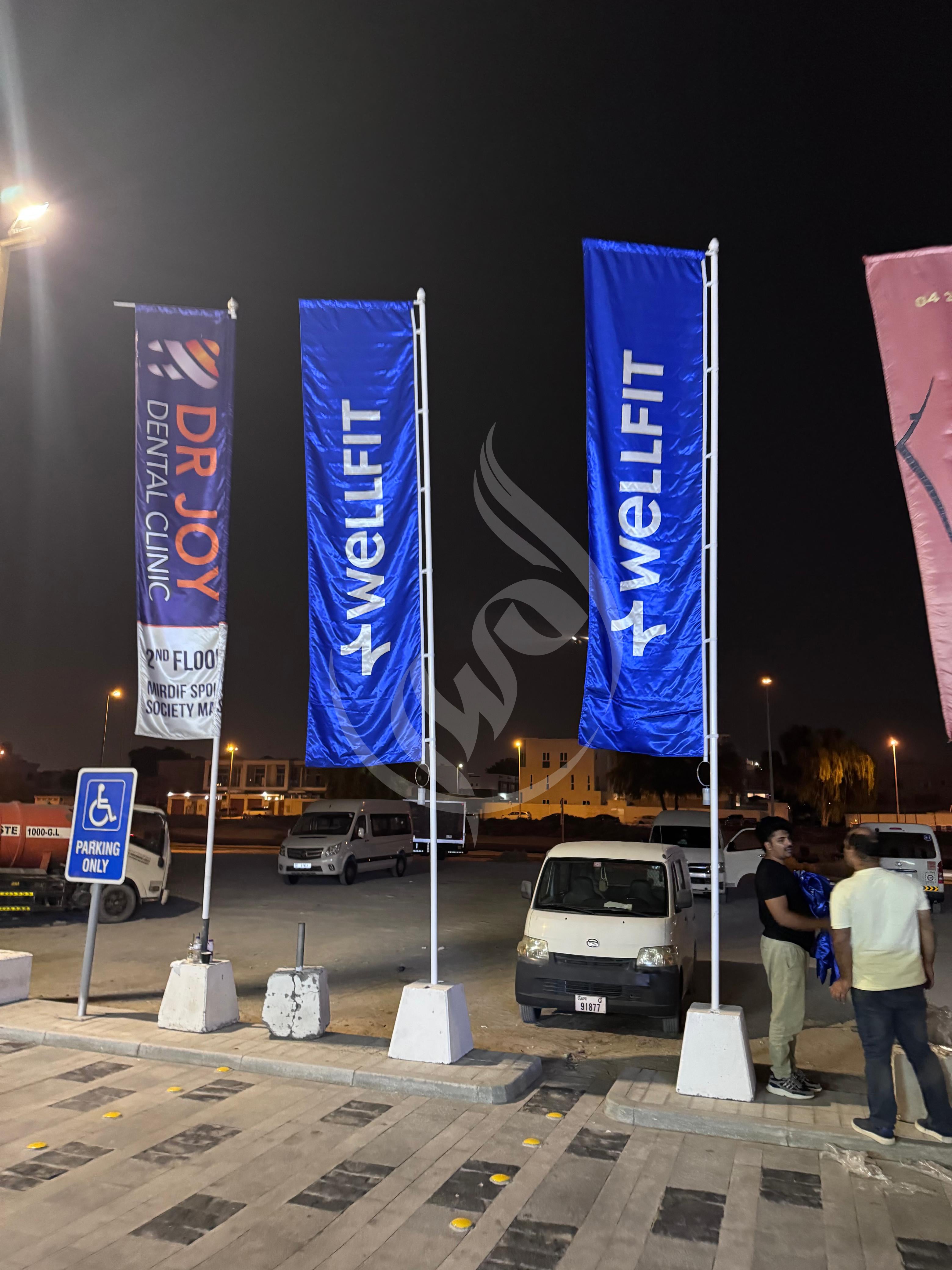 Three blue flags with 'Dr. Joy', 'Wellfit', and 'Dental Clinic' branding in an outdoor setting at night.