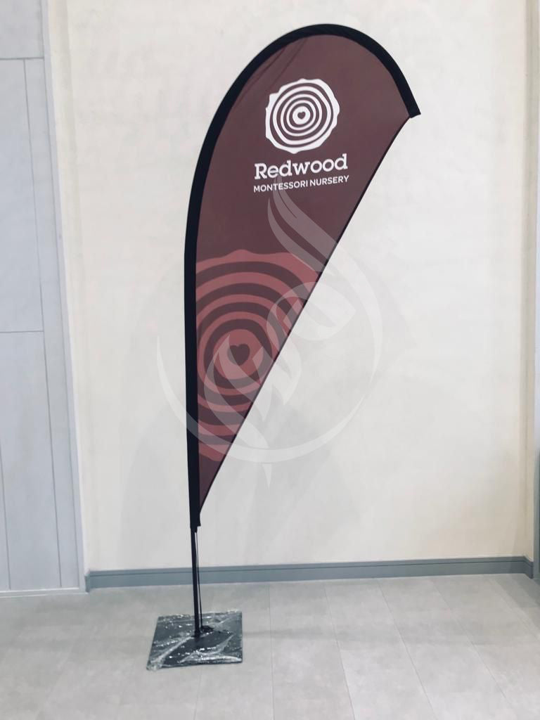 Teardrop flag with 'Redwood Montessori Nursery' logo against a plain wall.